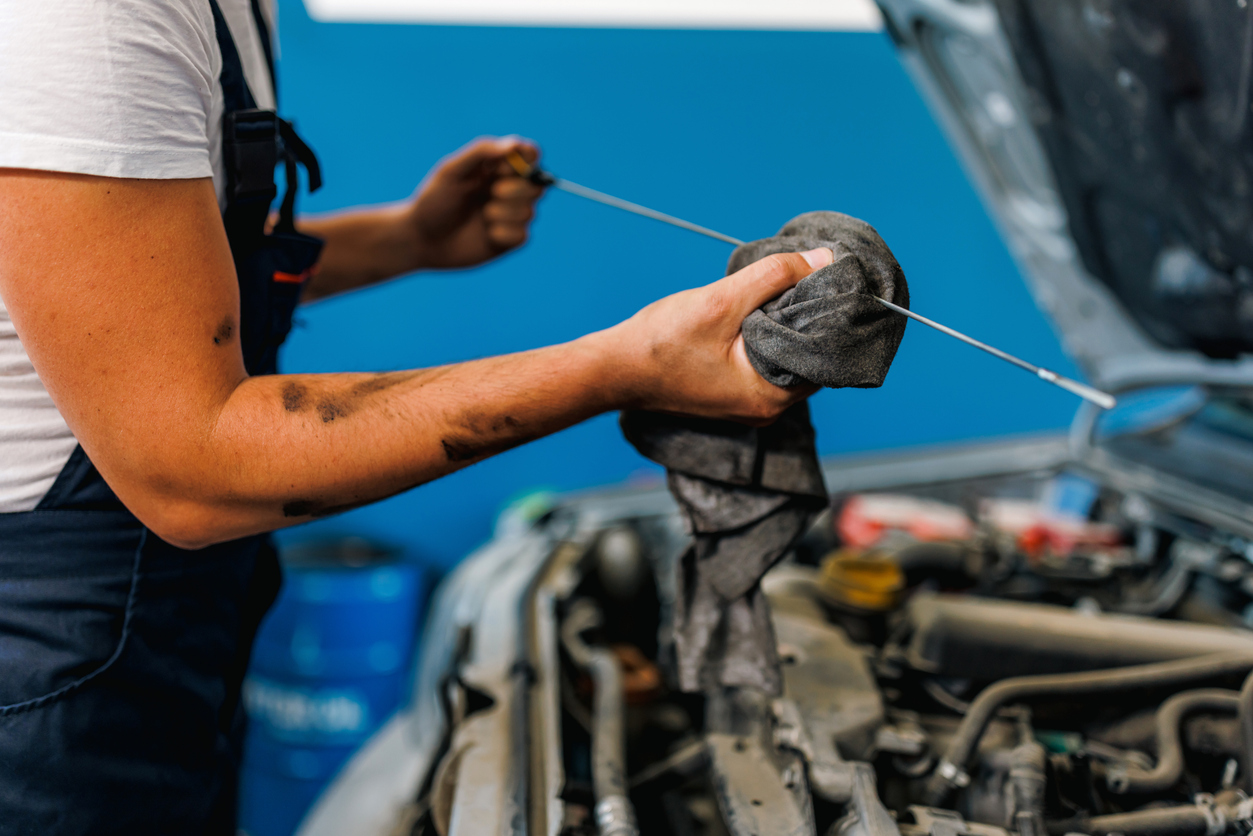 A close-up picture of a young Caucasian male mechanic standing next to a car with a popped hood and wiping a car part from motor oil, using a rug.
